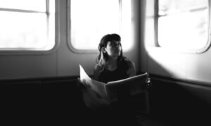 A photo of a woman sitting on a bench with an open newspaper in her hands as she looks out a nearby window. This is to demonstrate a journalist traveling somewhere new for freelancer conference season