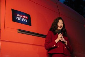 A stock image of a broadcast journalist holding a microphone and smiling. She is standing in front of a "breaking news" sign. This is to demonstrate journalistic objectivity.