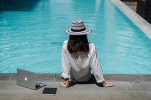 A travel blogger sitting at the side of the pool with her back facing the camera. A notebook and a laptop are beside her to illustrate Getting Invited on Press Trips